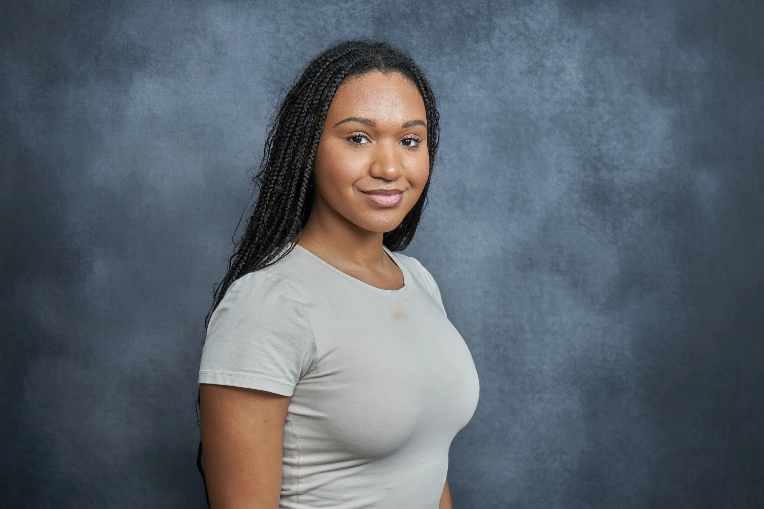 A young woman with long braided hair, wearing a light-colored t-shirt, standing against a dark background and smiling softly.