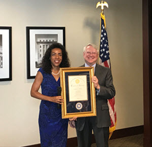 A woman and a man standing indoors, both smiling, holding a framed certificate or award. The woman is wearing a blue dress and the man is in a suit. An American flag is in the background.