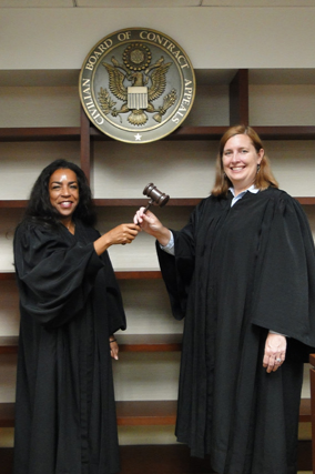 Two women in judicial robes exchanging a gavel in front of a seal that reads 'Board of Contract Appeals.'