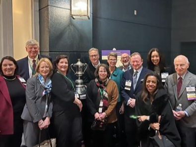 A group of people at an indoor event, some holding awards, standing in front of a dark wall with a blue plaque, smiling for the photo.