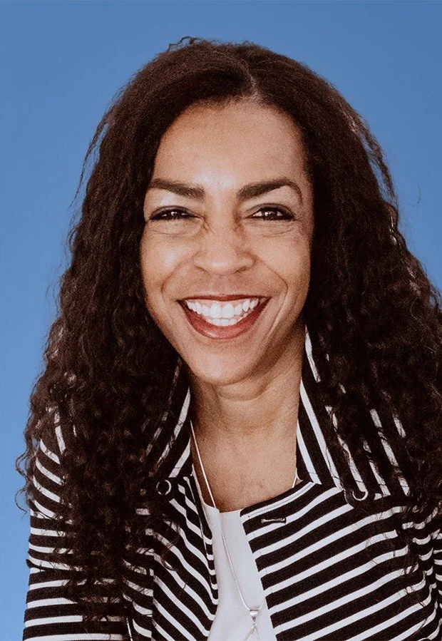 A woman with long, curly dark hair, smiling broadly against a blue background. She is wearing a black and white striped shirt and a necklace.