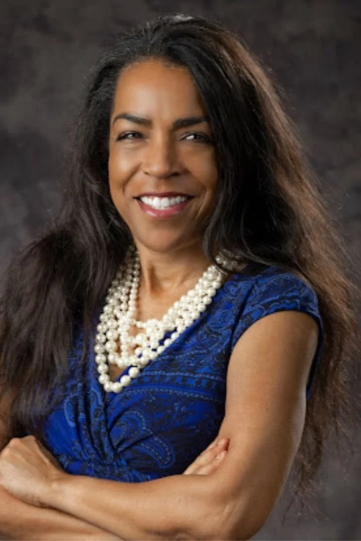 Woman with long dark hair wearing a blue patterned dress and multiple strands of pearls, smiling with arms crossed against a dark background.