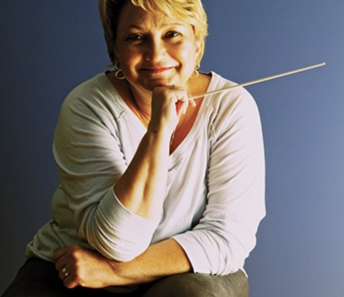 Portrait of a smiling woman with short blonde hair, wearing a white top, holding a conductor's baton, against a blue background.