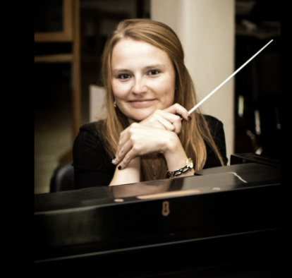 A young woman with red hair smiling and resting her chin on her hands behind a piano