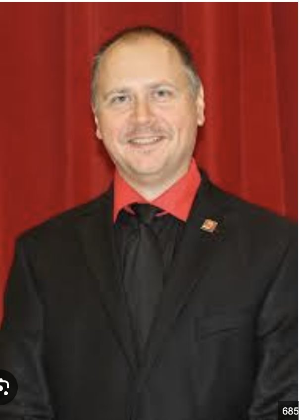 A man in a black suit with a red shirt and black tie, standing in front of a red curtain.