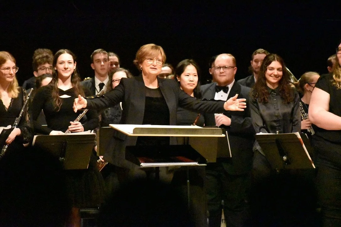 Conductor leading a large choir or orchestra on stage with music stands, black backdrop, and performers in formal attire.
