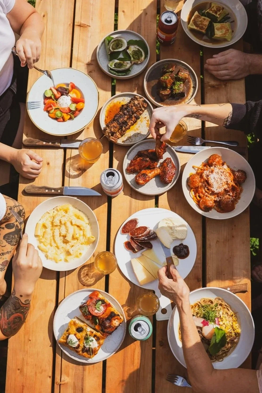 Repas partagé avec diverses assiettes de plats comme pâtes, pizza, viande, légumes, fromage, et boissons, sur une table en bois.