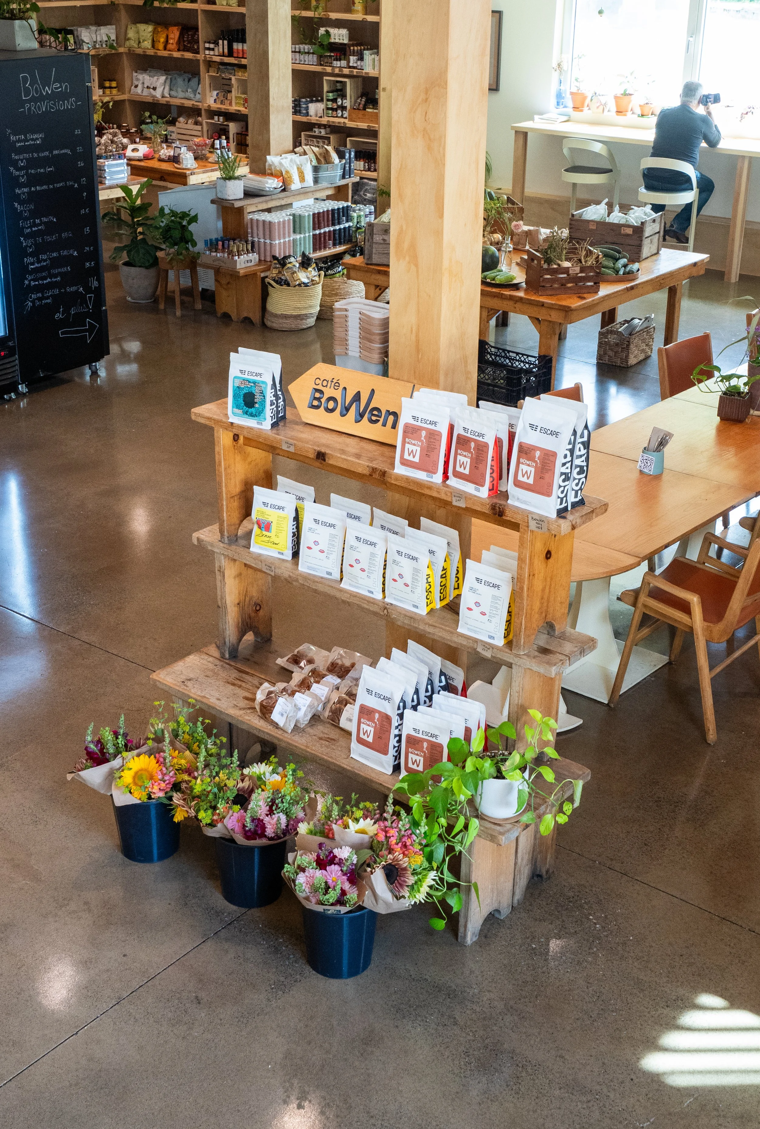 Une boutique ou marché avec étagères en bois contenant des produits divers, un tableau noir avec une liste, plusieurs paniers de fleurs colorées et des sacs de café sur une table en bois.