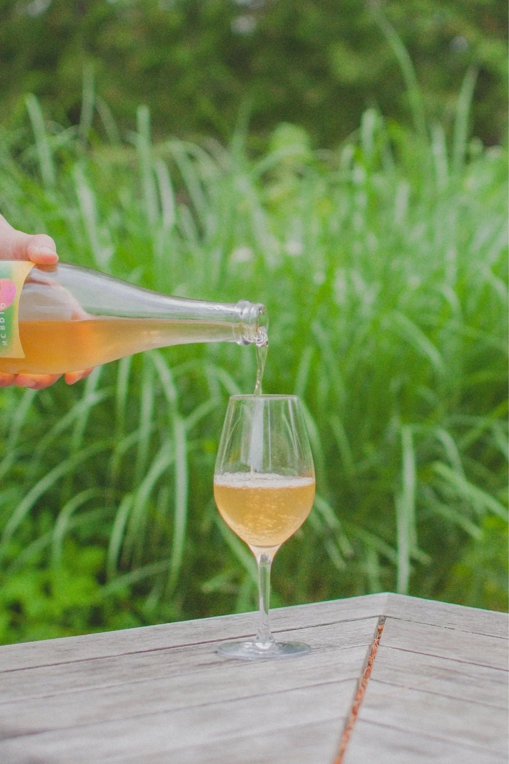 Une personne verse une boisson dorée dans un verre à vin dans un environnement extérieur avec des plantes vertes en arrière-plan.