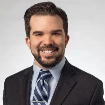 A young man with dark hair and a beard, smiling, wearing a suit and tie, posing against a plain light gray background.