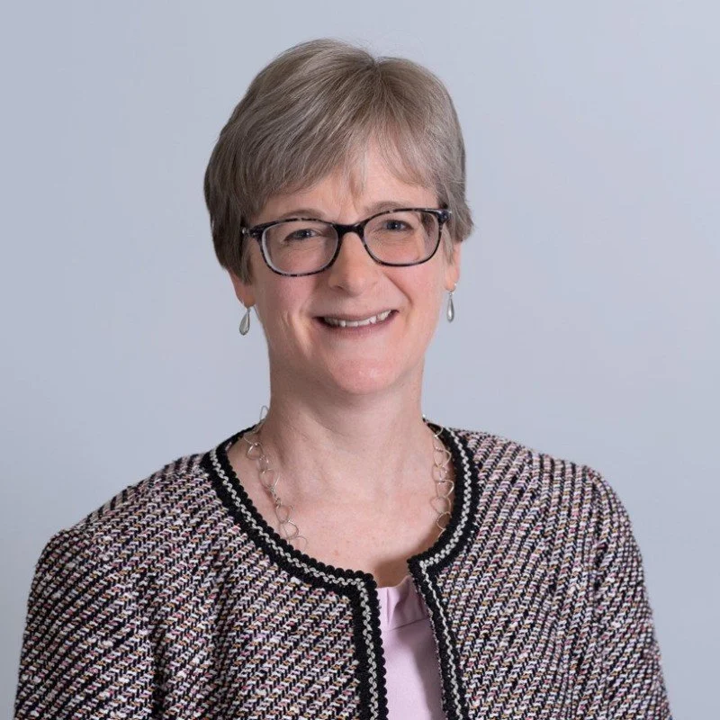 A portrait of a smiling woman with short gray hair, glasses, earrings, and a patterned jacket against a light blue background.