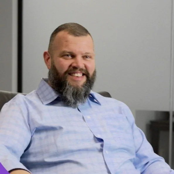 A smiling man with a beard and short hair sitting in an office, wearing a light blue checkered shirt.