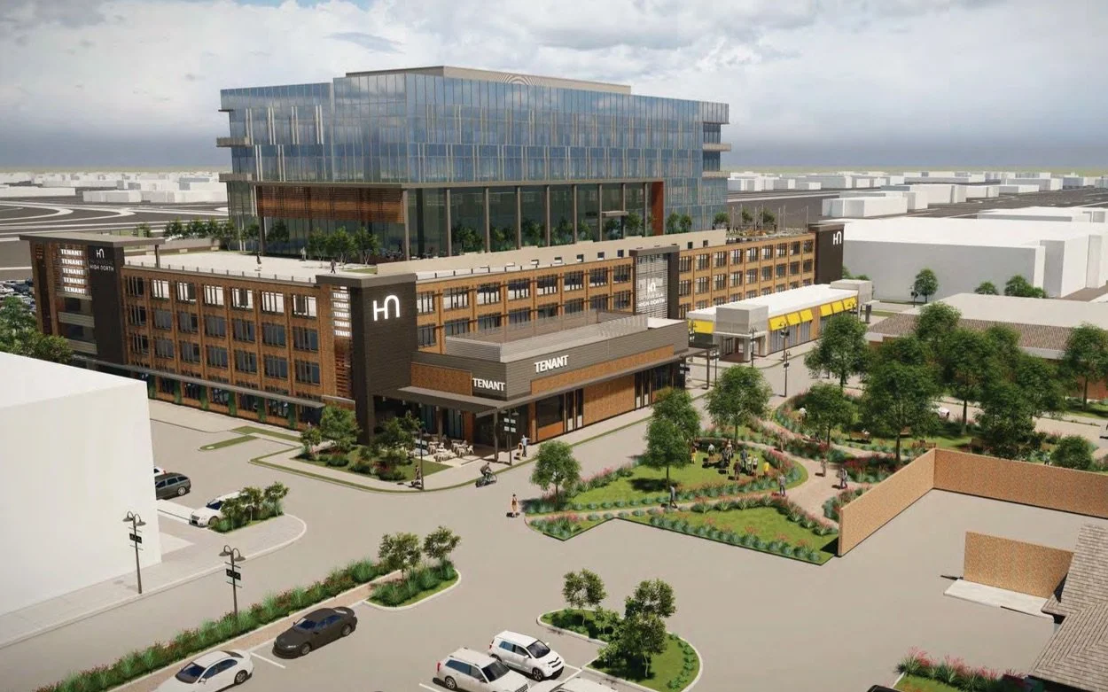 A modern multi-story building with retail shops on the ground floor and office or hotel space above, surrounded by parking lots, greenery, and a landscaped park.