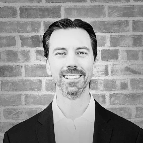 A black-and-white photo of a man with dark hair slicked back, a beard, and mustache, wearing a suit jacket and white shirt, standing in front of a brick wall.