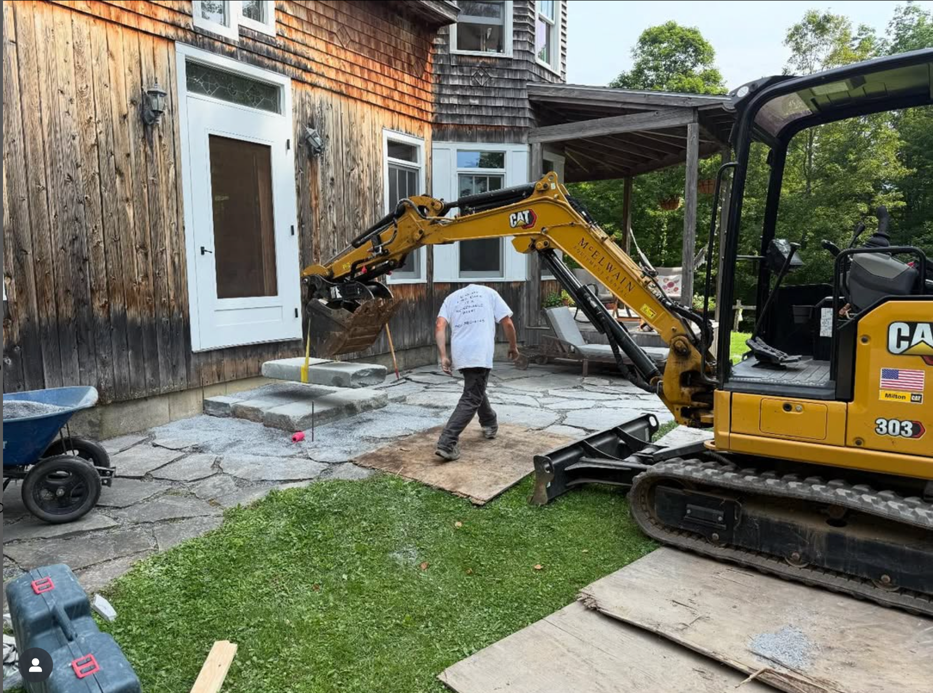 A person working on outdoor patio construction with a yellow CAT excavator lifting concrete blocks, in front of a wooden house with a door and windows, surrounded by greenery.