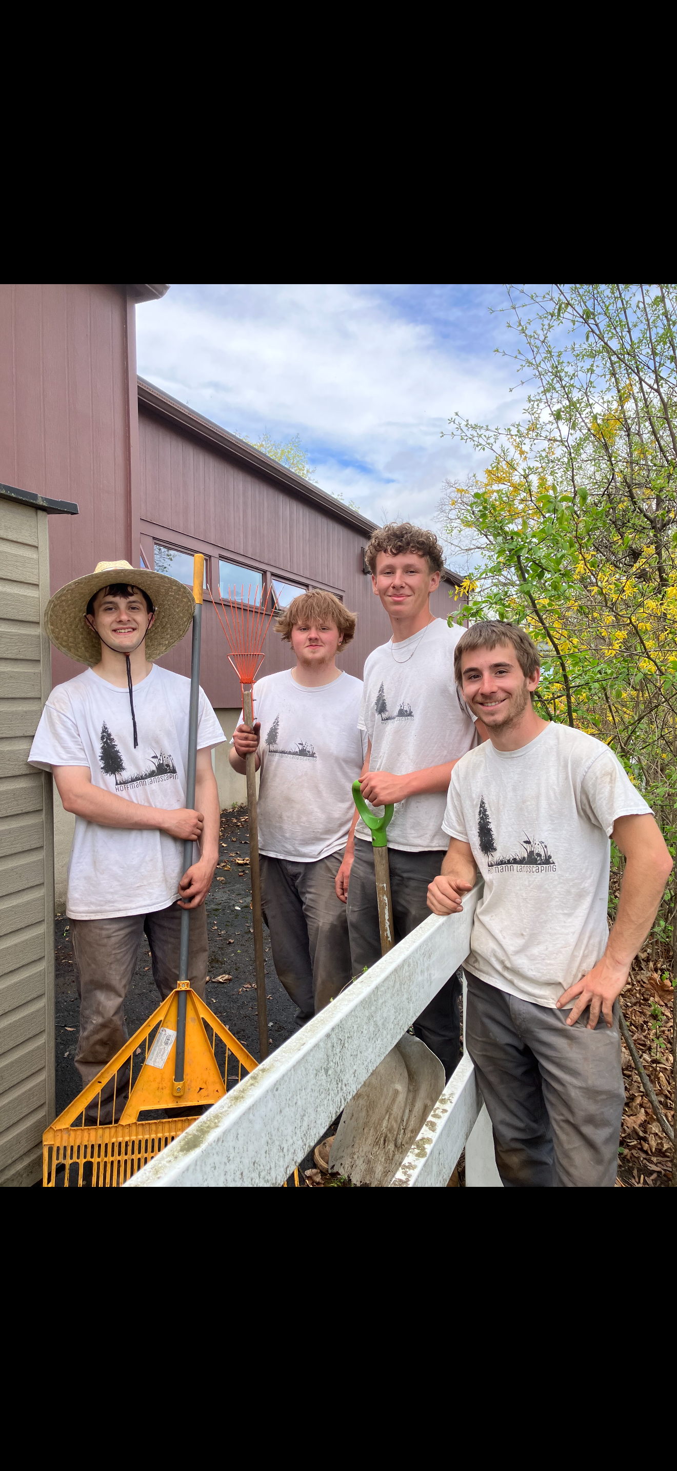 Group of four young men standing outdoors, holding gardening tools, smiling, with a house and trees in the background.