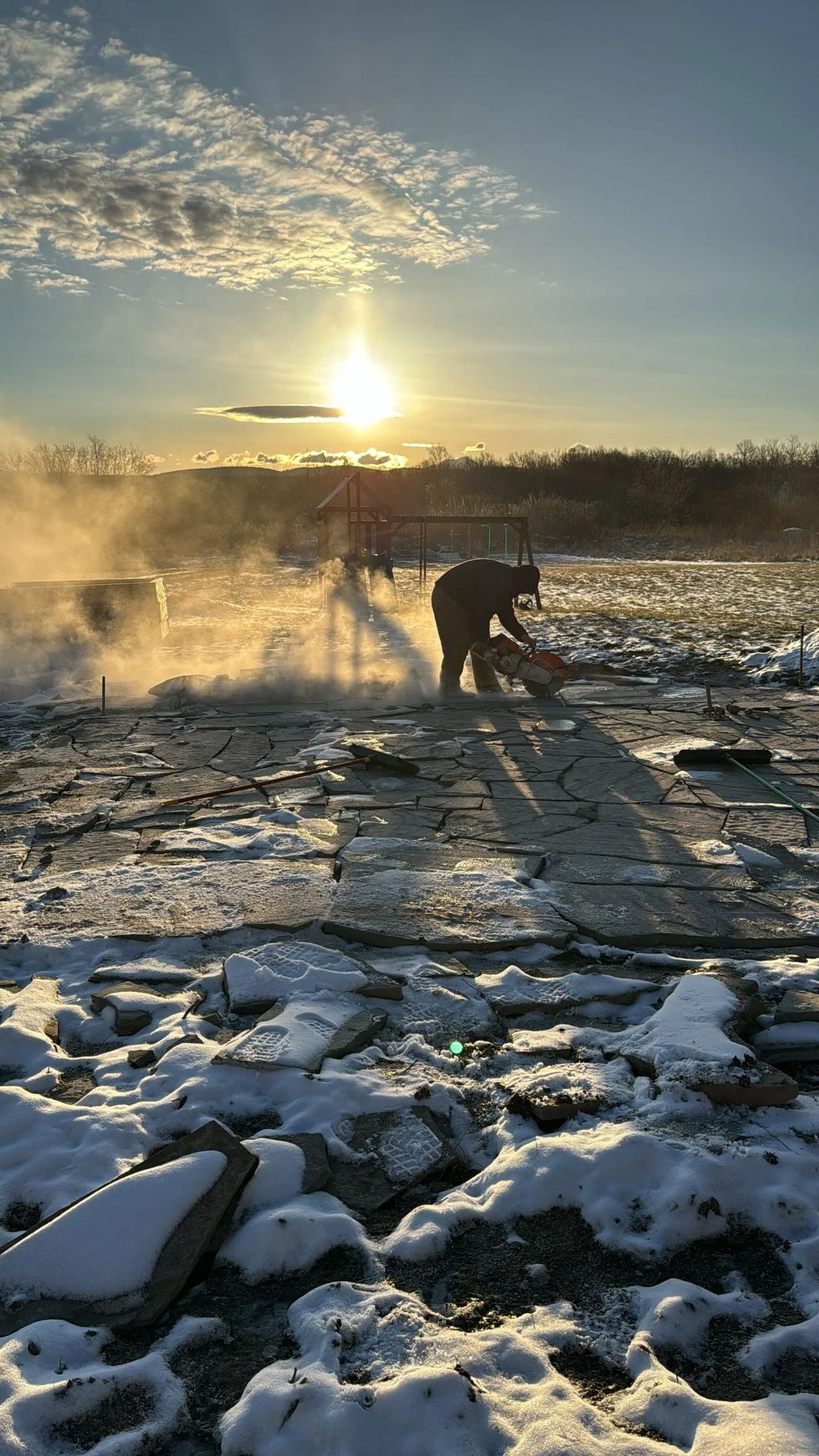 A person using a chainsaw on a stone patio outside, with snow and ice on the ground, during sunset or sunrise.