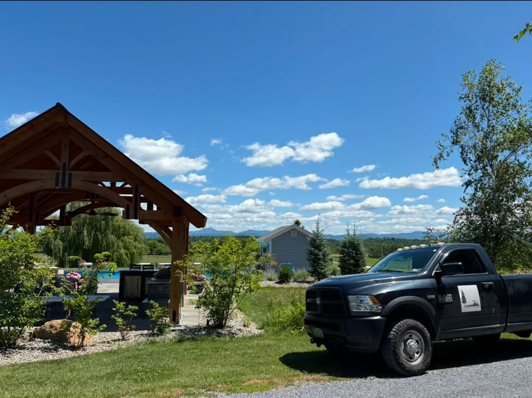 A black pickup truck parked on a gravel area beside a grassy lawn, with a wooden pavilion, trees, and a blue house in the background under a partly cloudy sky.