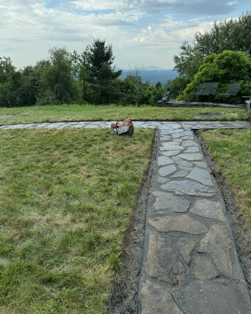 A stone pathway being installed on a grassy yard with a landscape of trees and hills in the background. There is a chainsaw on the ground and two empty benches with a Buddha statue nearby.