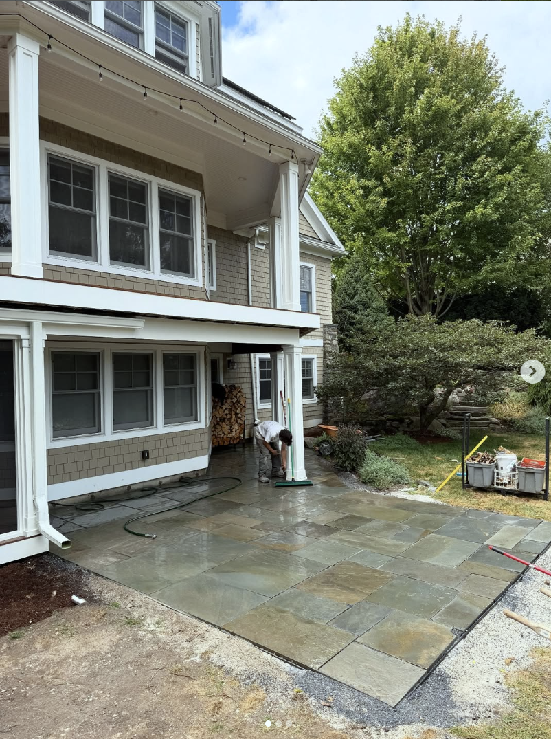 A person installing or cleaning a stone patio outside a house with a covered porch, multiple windows, and a pile of firewood. The yard has trees and bushes, with some construction tools and materials nearby.