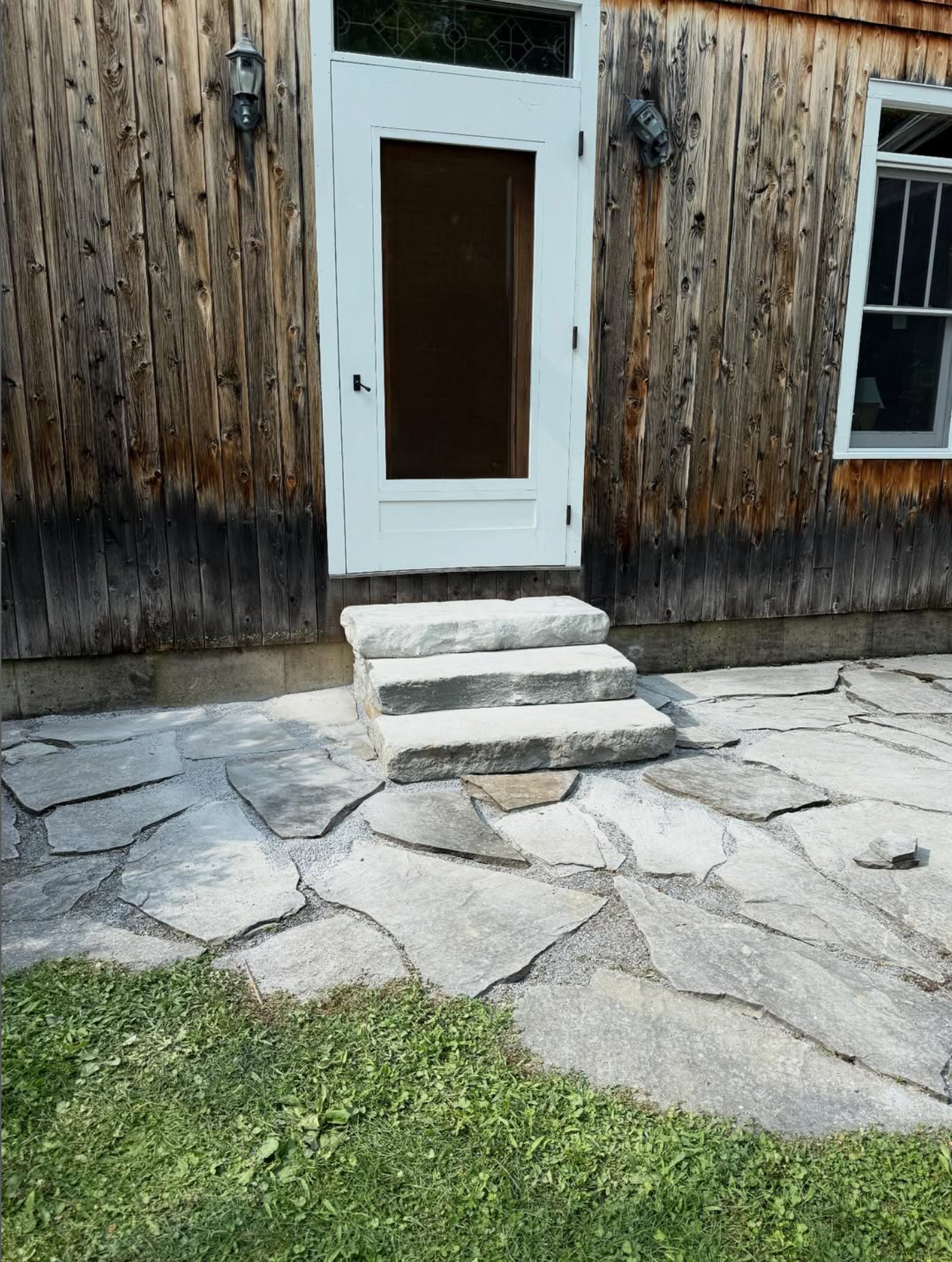 Stone steps leading to a wooden door on a house with weathered wood siding, a window, and a flagstone patio.