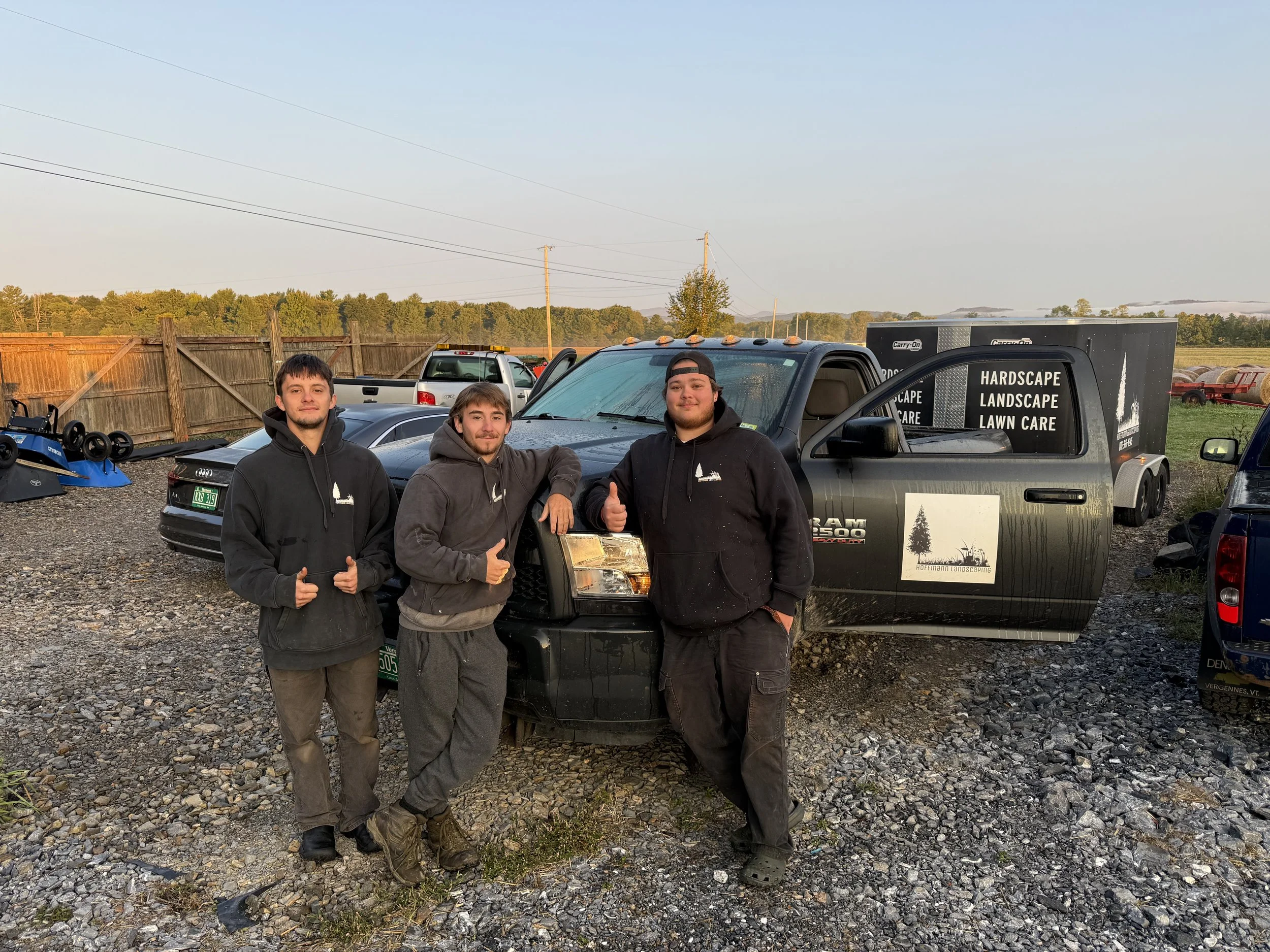 Three men standing in front of trucks and a car in a gravel lot, with a wooden fence and trees in the background, during sunset.