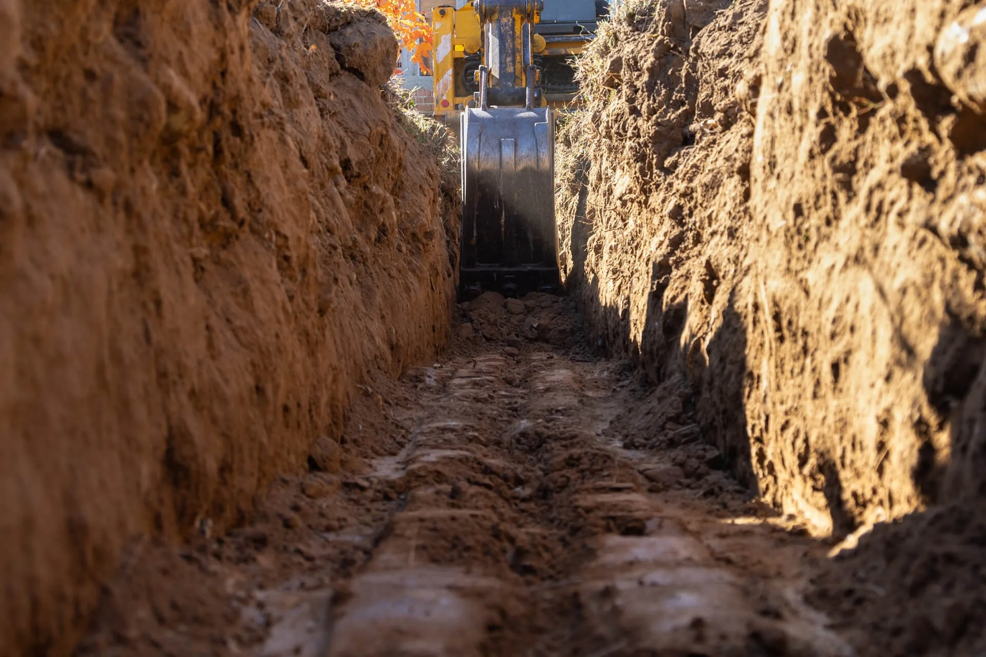 Close-up of a construction excavator bucket digging a trench in brown soil with a brick building and orange leaves in the background.
