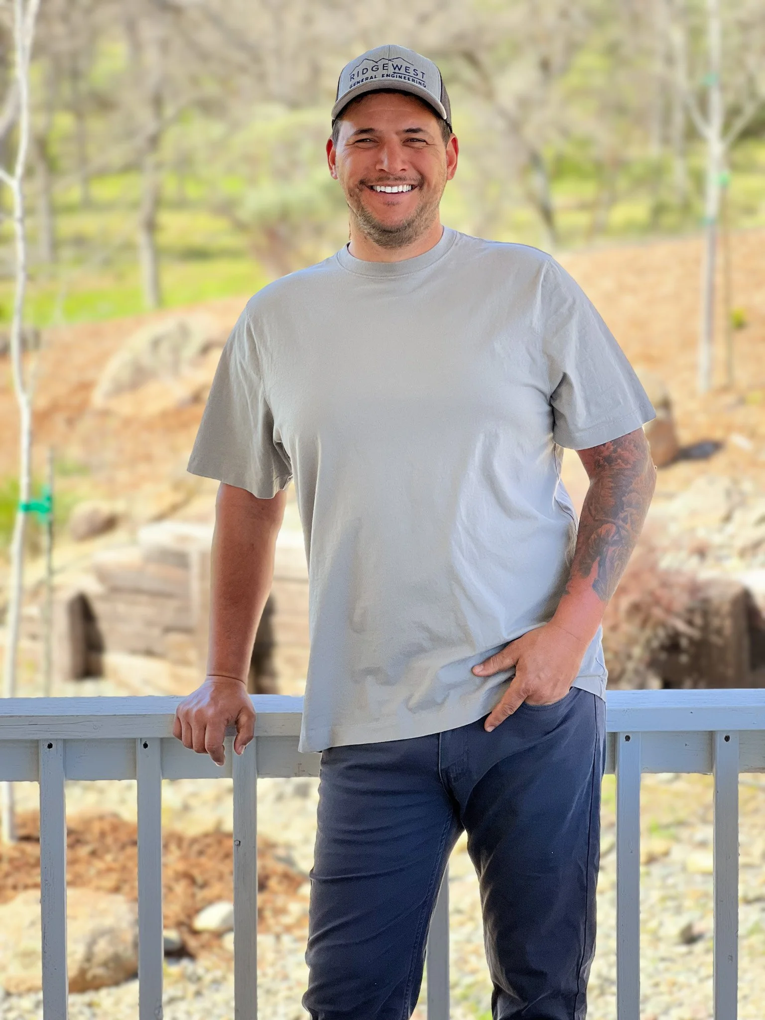 A smiling man wearing a light gray T-shirt, dark pants, and a gray cap that reads 'RIDGEWEST GENERAL ENGINEERING' stands outdoors leaning on a white railing with a blurred natural background of trees and rocks.