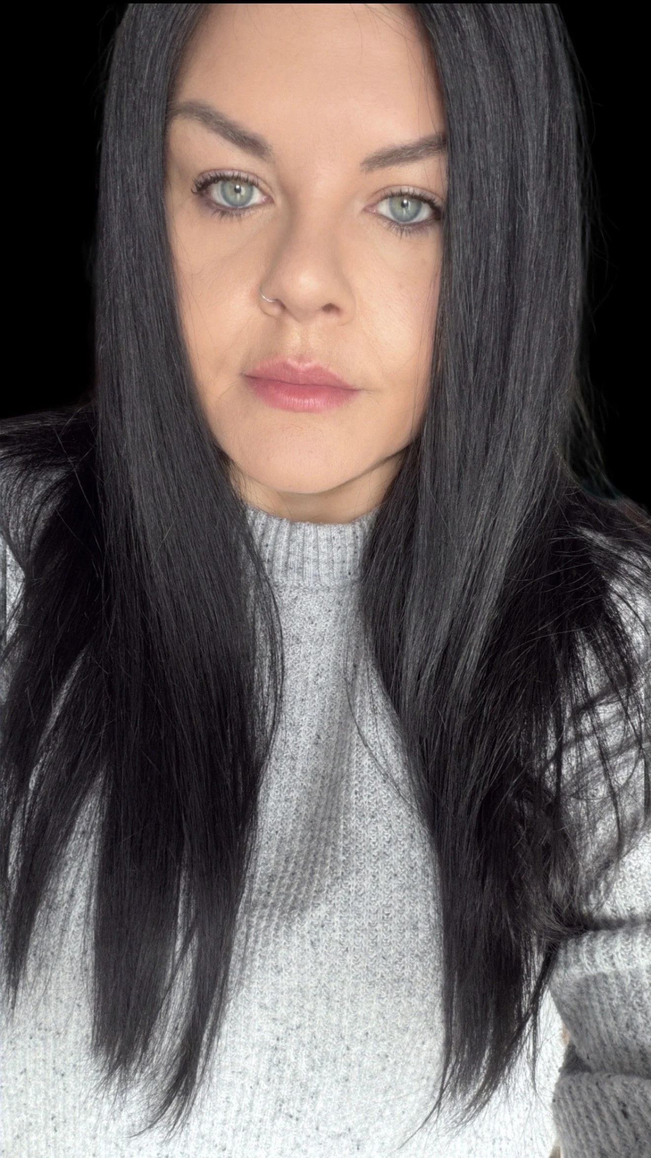 A young woman with long, wavy red hair and blue eyes sitting on a chair, smiling at the camera, indoors with a light gray wall and a white door in the background.