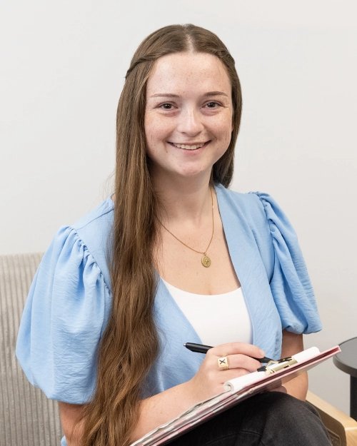 A man with curly brown hair, a beard, and a mustache sitting cross-legged on a dark-colored sofa in a white room, wearing a light-colored, striped shirt. There is a white pillow with colorful motivational messages behind him.