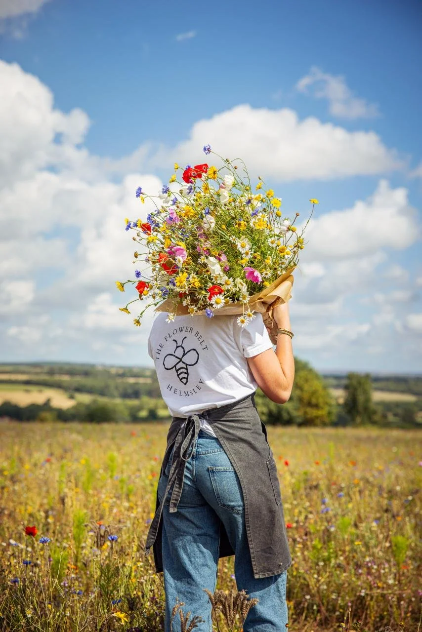 A person standing in a wildflower field holding a large bouquet of wildflowers over their head, facing away, wearing blue jeans, a white shirt, and a black apron, with a scenic landscape of fields and trees under a blue sky with clouds in the background.