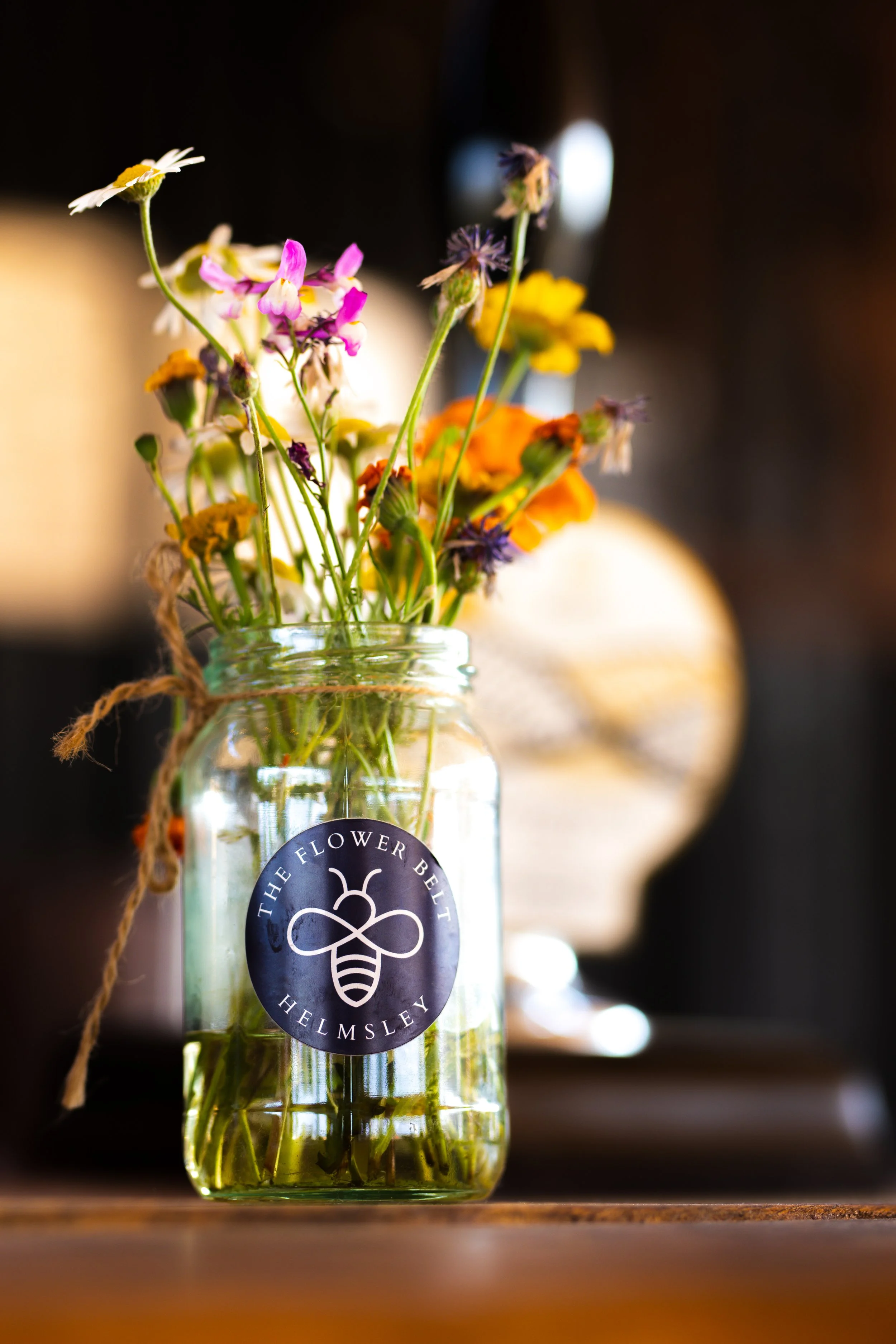 A glass jar filled with water and colourful wildflowers, sitting on a wooden surface. The jar has a label with The Flower Belt's logo, tied with a piece of twine. The background is blurred with warm and dark tones.
