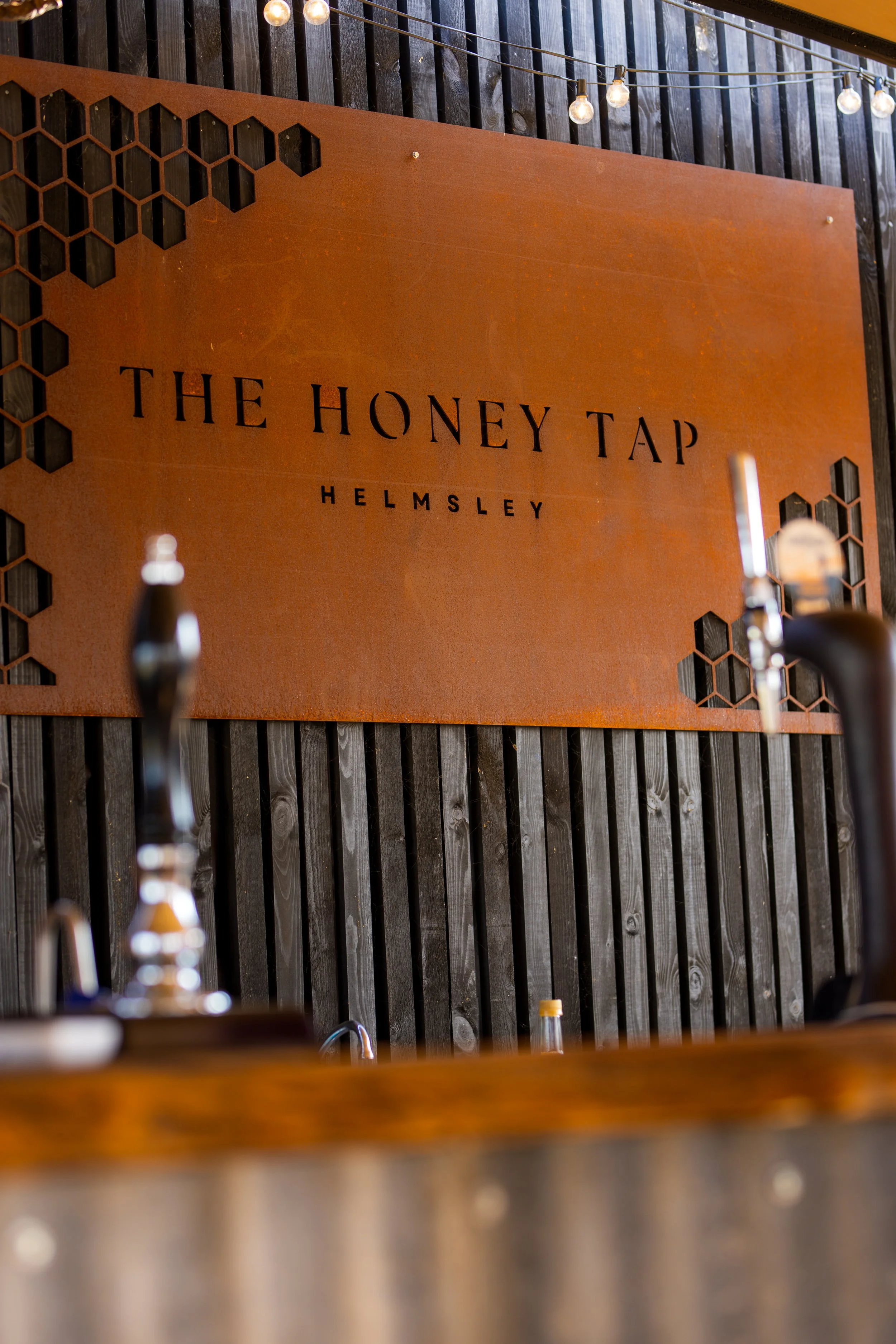 Sign reading 'The Honey Tap Helmsley' on a wooden wall with string lights above.