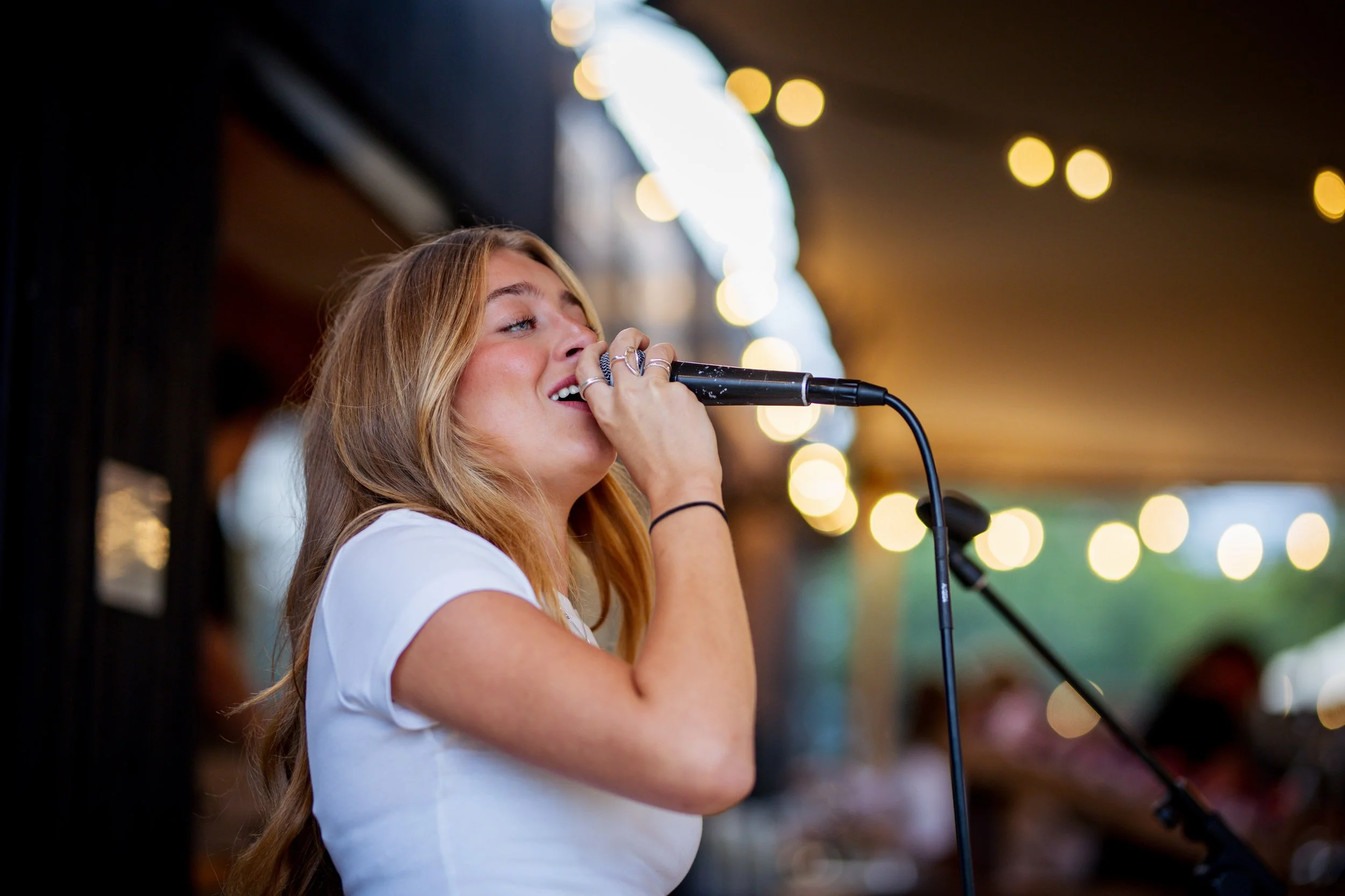 A young woman singing into a microphone at an outdoor event with festoon lights in the background.