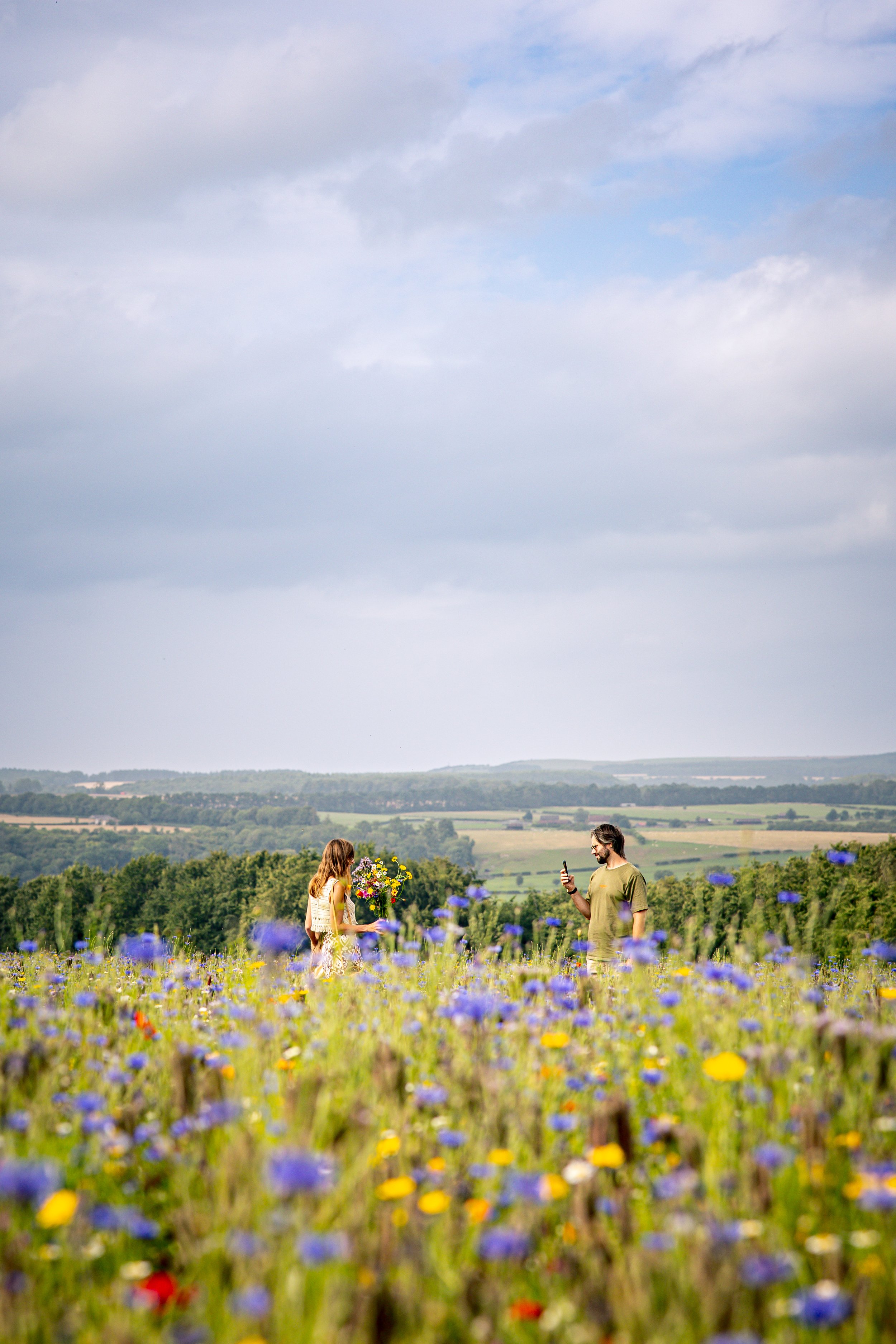 A man and woman in a field of colourful wildflowers, with rolling hills and a cloudy sky in the background. The woman is holding a bouquet of flowers and the man is taking a photo with his phone.