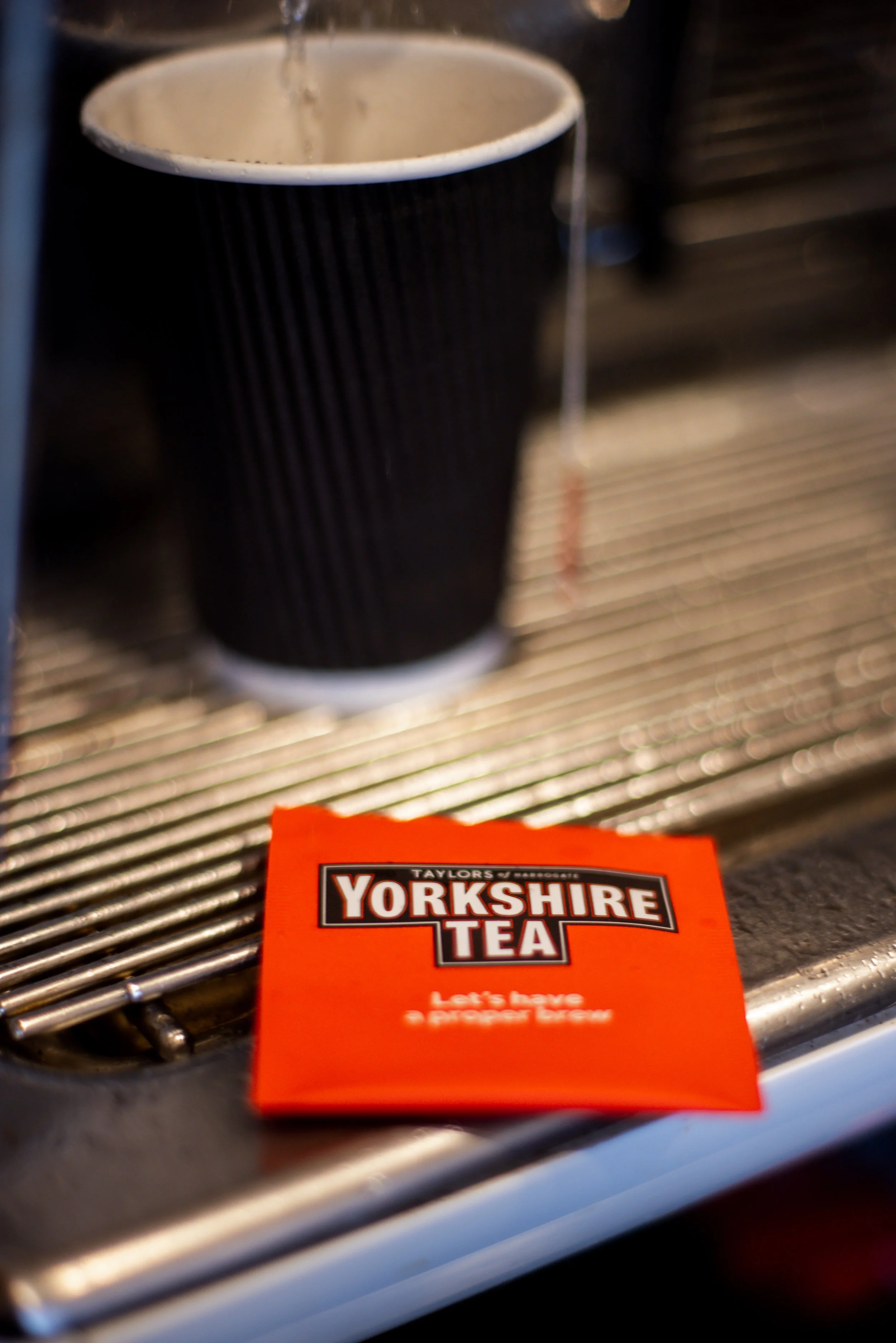 A black disposable cup being filled with water from a faucet. The cup is placed on a metal sink grate. In the foreground, there is a red packet of Yorkshire Tea with the text 'Let's have a Proper Brew'.