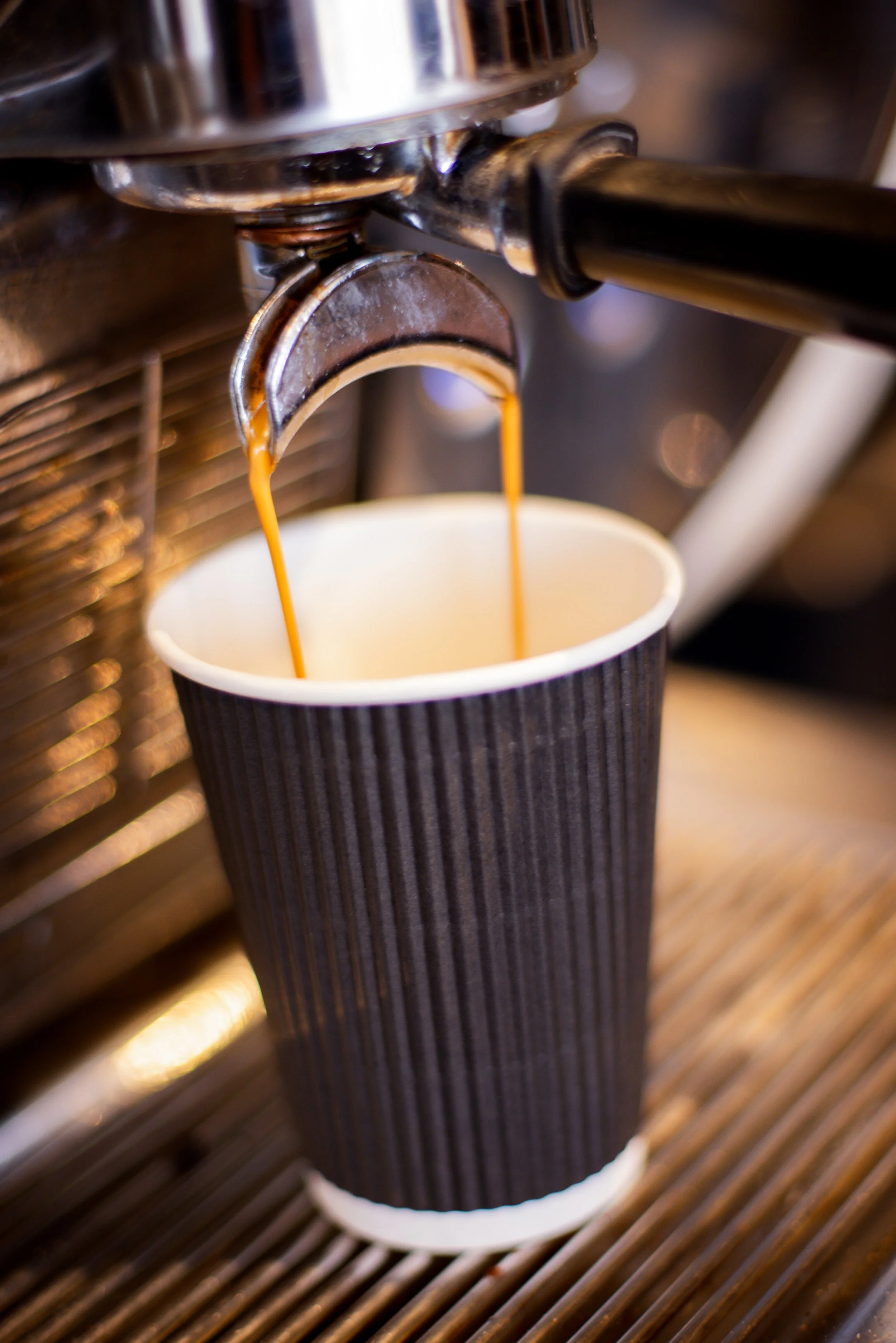Close-up of a coffee machine dispensing fresh espresso into a black paper cup with ridges, placed on a wooden surface.