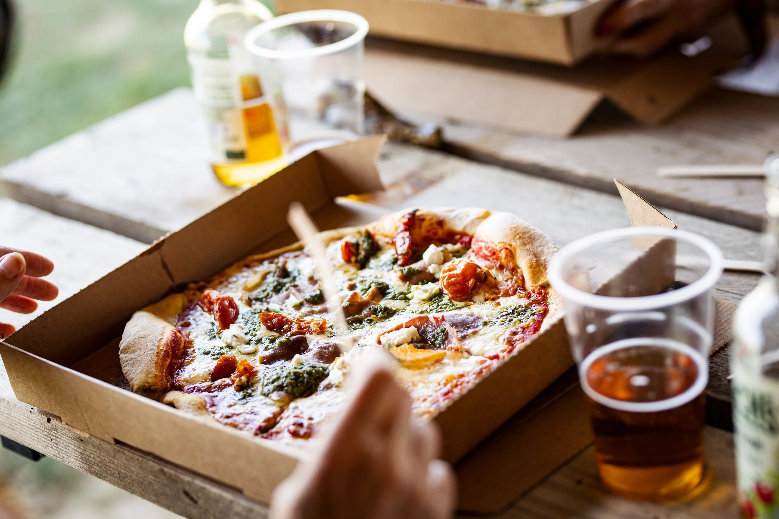 A person holding an open pizza box with a slice of pizza topped with vegetables and cheese. There are cup drinks and condiments on a wooden table.