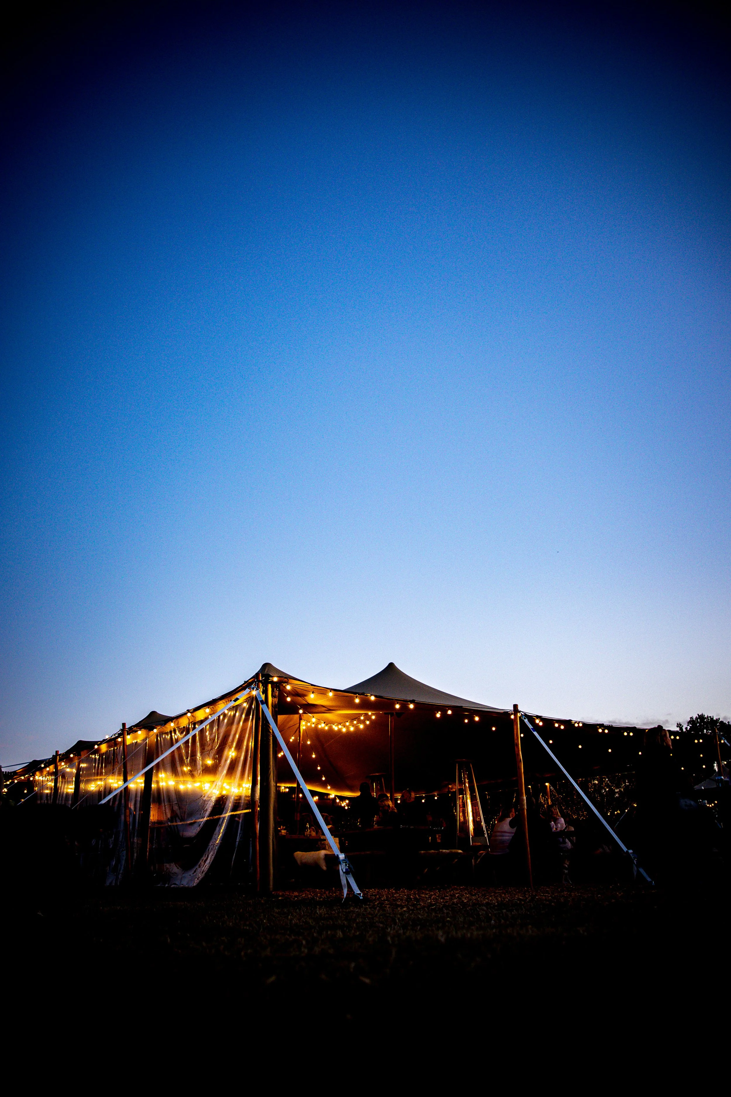 Outdoor event tent illuminated with string lights during dusk, with a darkening sky and some trees visible in the background.