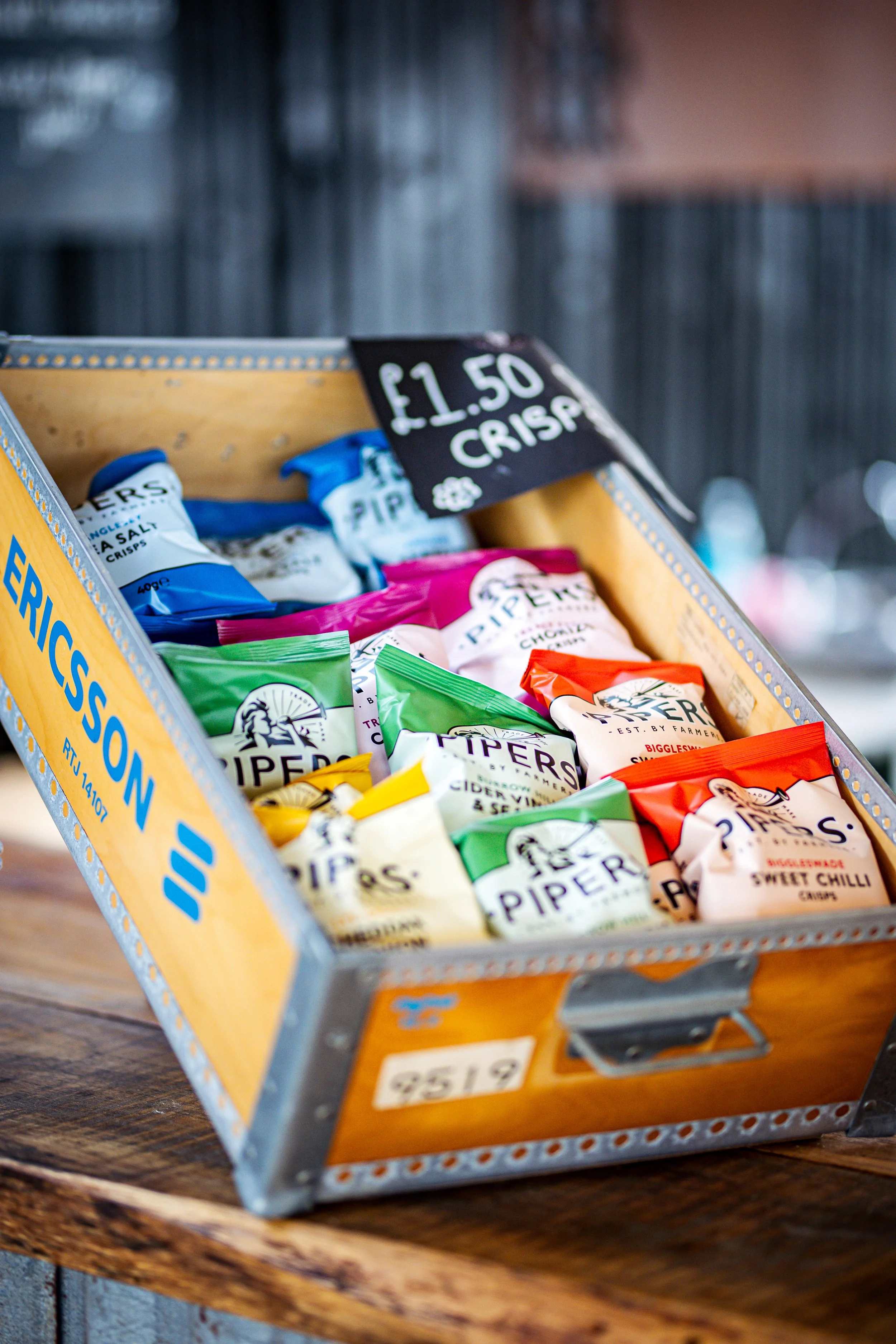 A display of assorted bagged crisps (potato chips) in a metal shopping basket with a sign indicating the price of £1.50 for each bag.