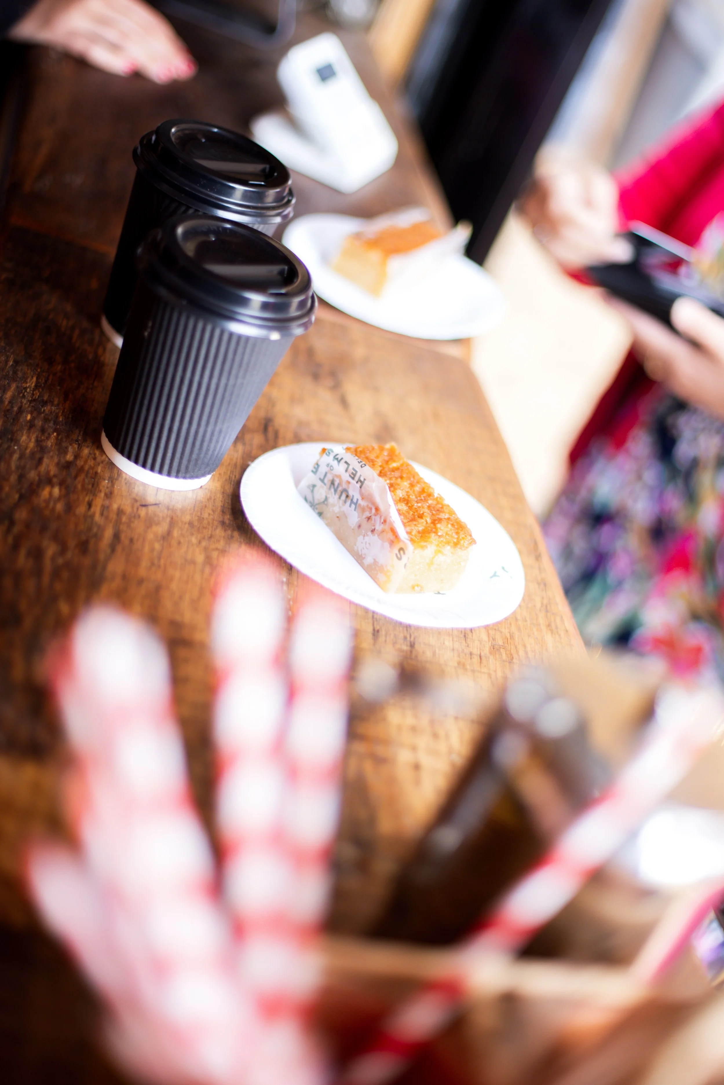 Three black to-go coffee cups on a wooden table with a person's hand and a plate of cake in the background.