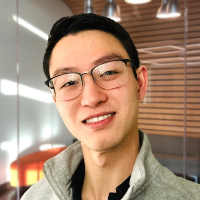 Young person wearing glasses and a gray jacket, smiling, in a modern office setting with wooden paneling and soft lighting.