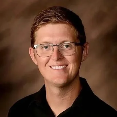 A young man wearing glasses and a striped shirt smiling outdoors with a hazy background.