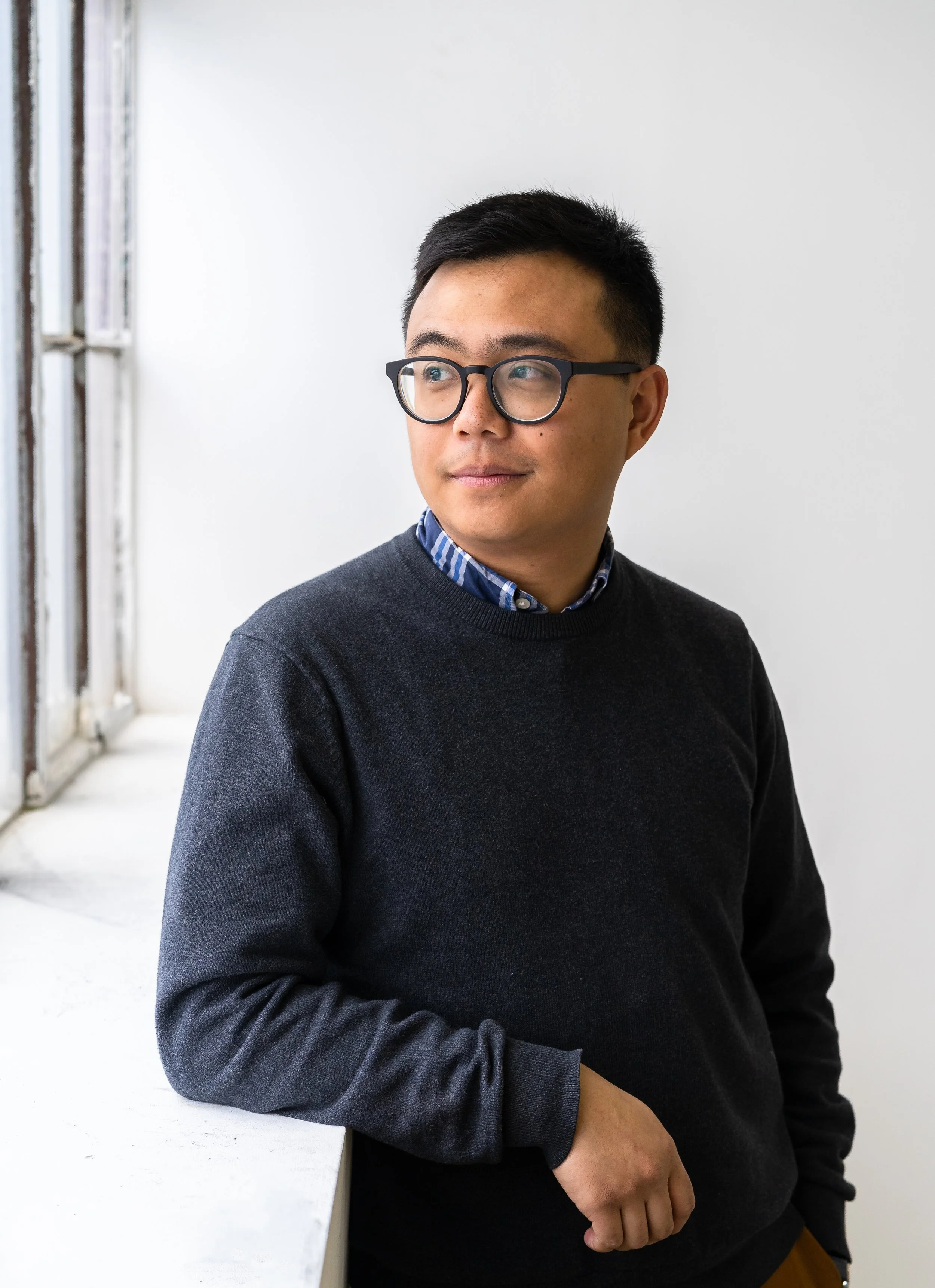 Man wearing glasses and a dark sweater, standing by a window, looking to the side.