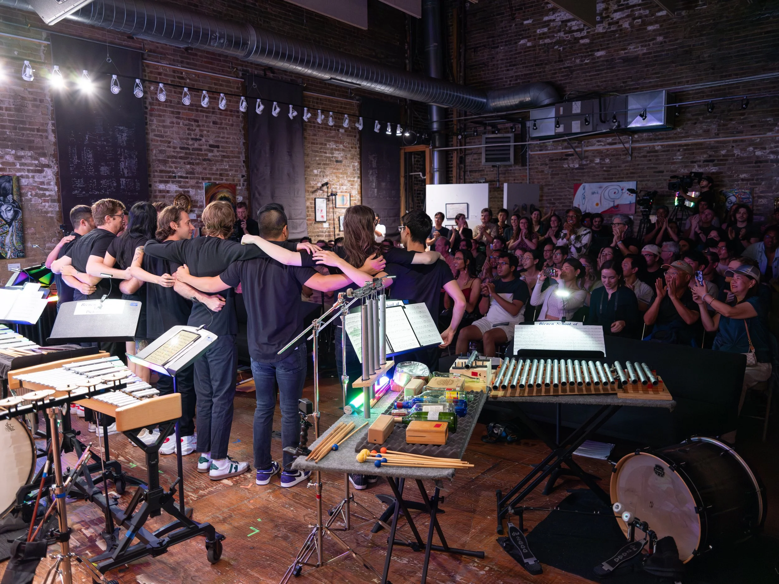 Musicians on stage in a brick-walled venue, standing in a line with arms around each other, facing an applauding audience. Percussion instruments and music stands are in the foreground.