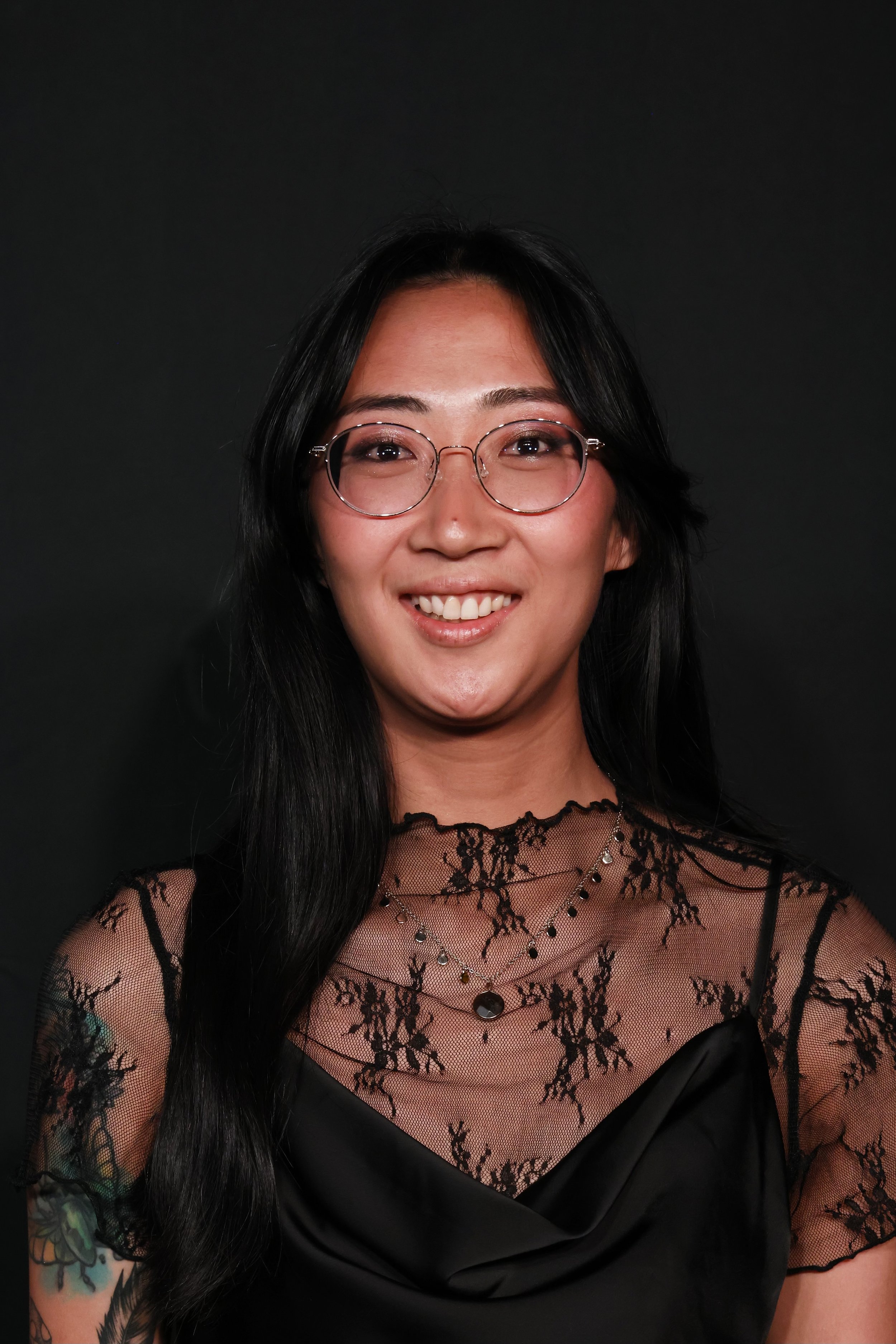 A person with long dark hair is smiling, wearing round glasses and a black lace top against a dark background.