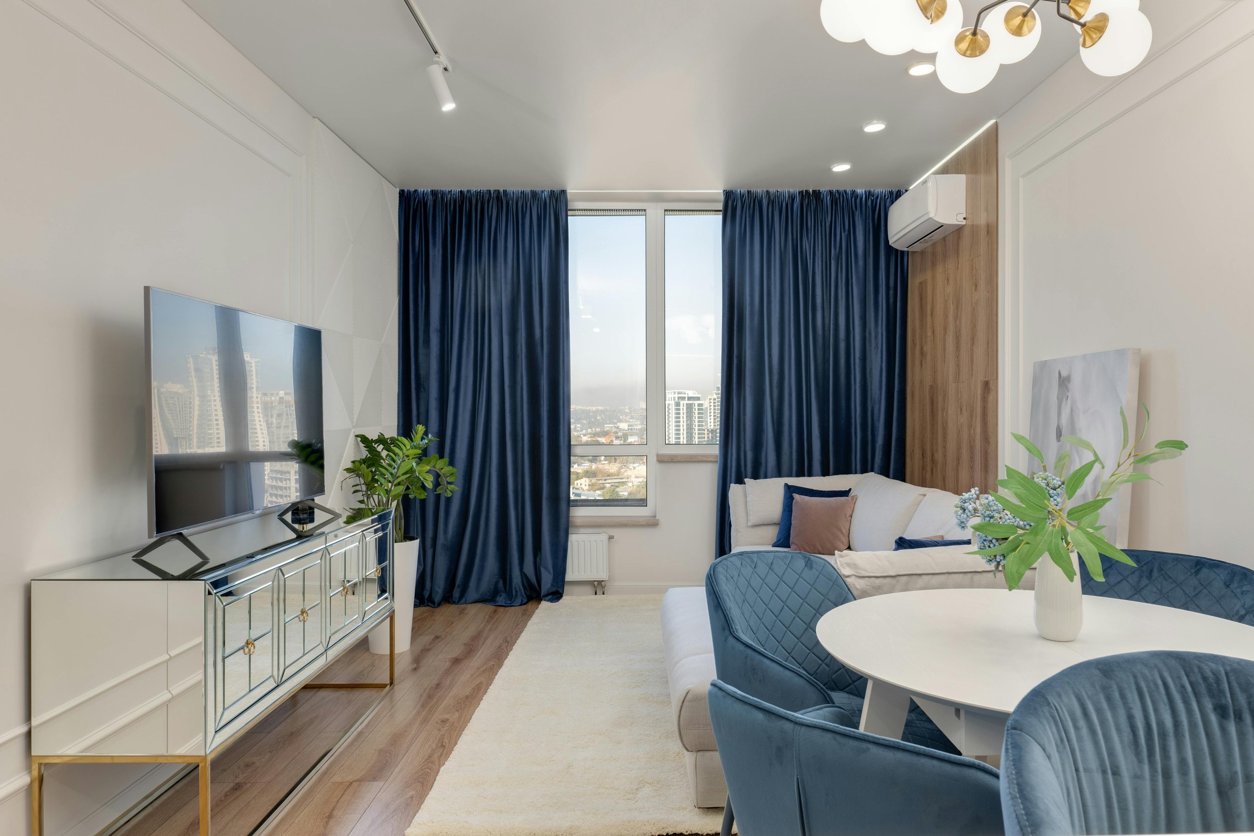 Modern living room with a white rug, blue curtains, and a light-colored sofa. A TV is mounted on a mirrored cabinet, with potted plants nearby. The room features blue chairs around a white round table, and a decorative light fixture on the ceiling.