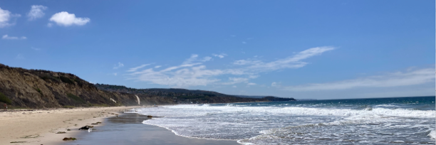 Biofield Tuning can help you feel great. A wide view of a sandy beach with waves crashing, cliffs in the distance, and a blue sky with some clouds.