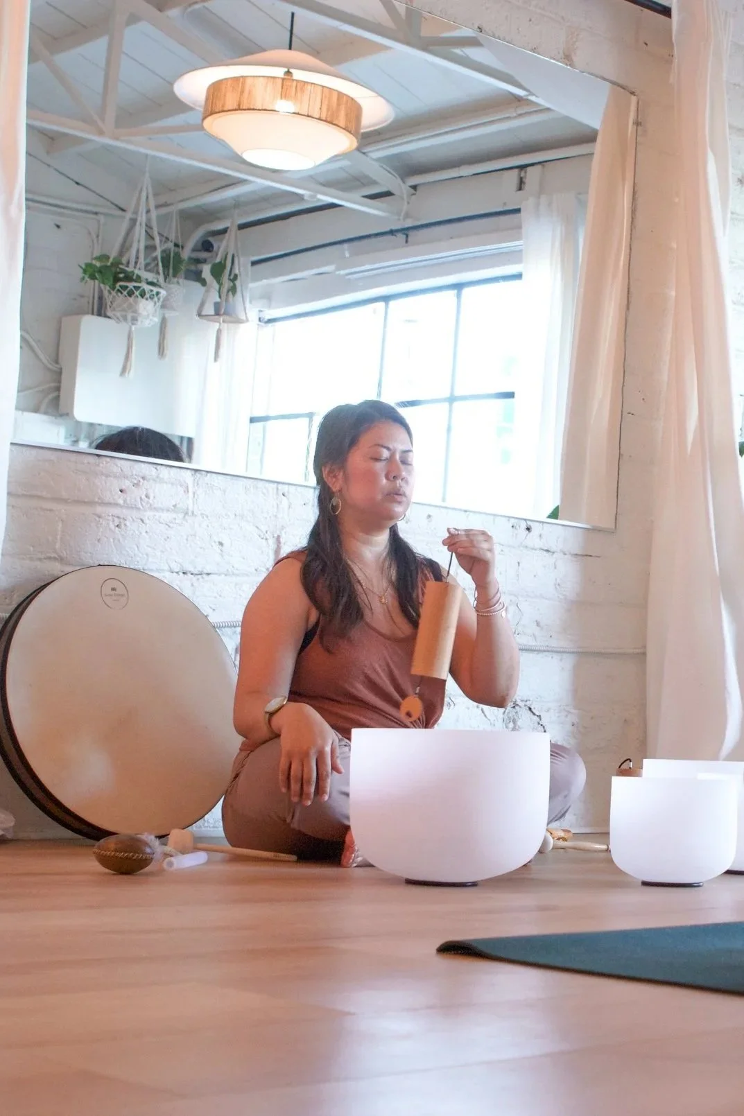A woman sitting cross-legged on a wooden floor, playing crystal singing bowls and holding a mallet in one hand. She has dark hair, is wearing a brown tank top, and has several pieces of jewelry. There is a large mirror on the wall behind her reflecting a bright window, white curtains, and hanging plants. A drum and other small percussion instruments are on the floor beside her.