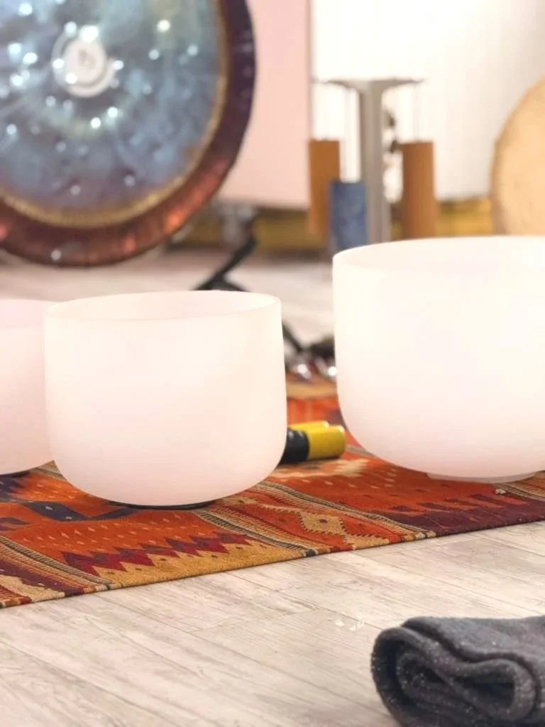 Three white crystal singing bowls on a colorful table runner, with a dark-colored cloth in the foreground and a clock in the background.